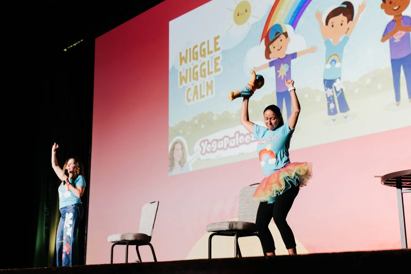 Two Yogapalooza presenters leading a movement song on stage at Frog Street Splash, one holding the puppy plush